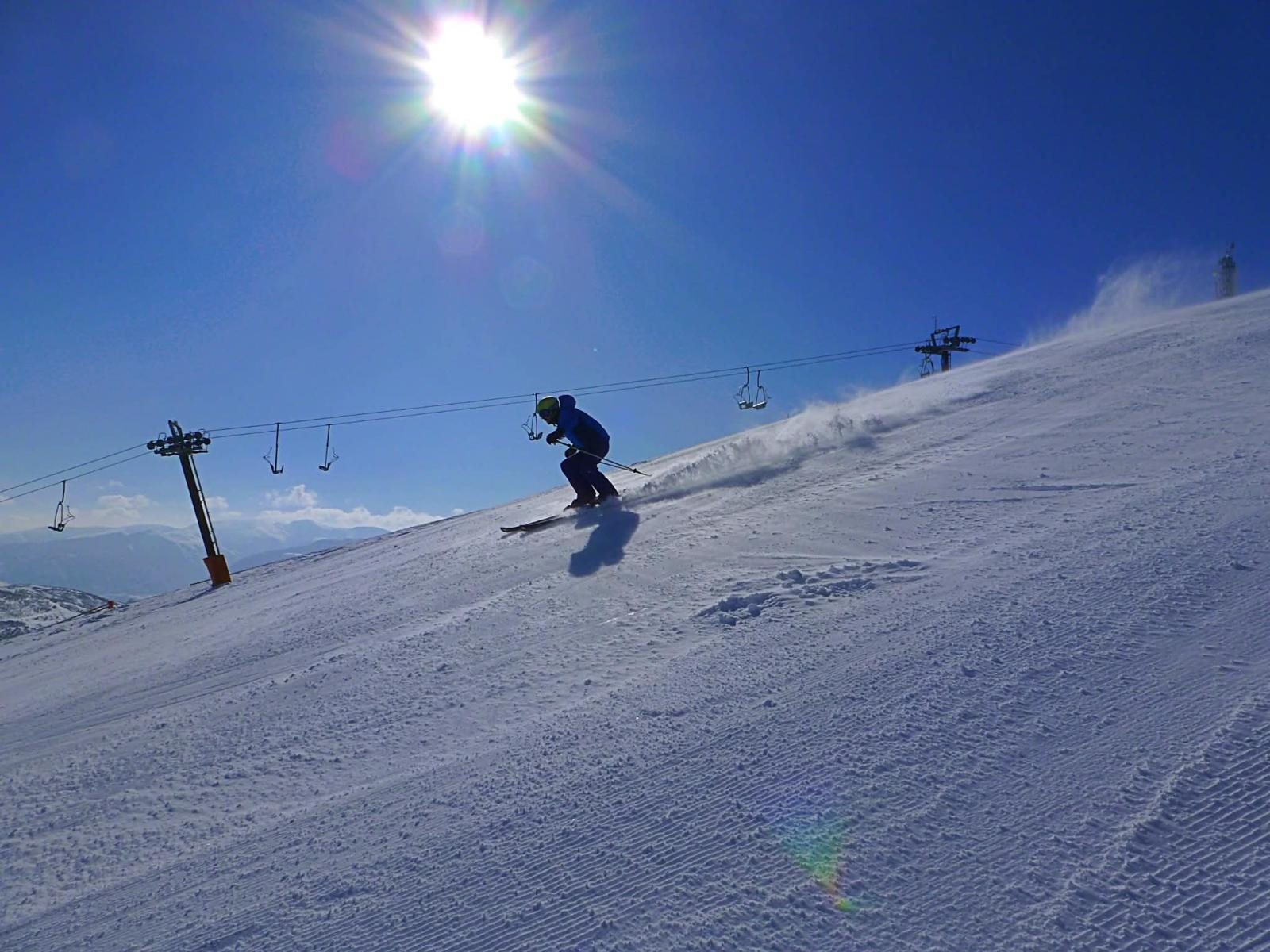 Un esquiador baja la pista a contraluz, levantando nieve, con el telesilla y el sol detrás